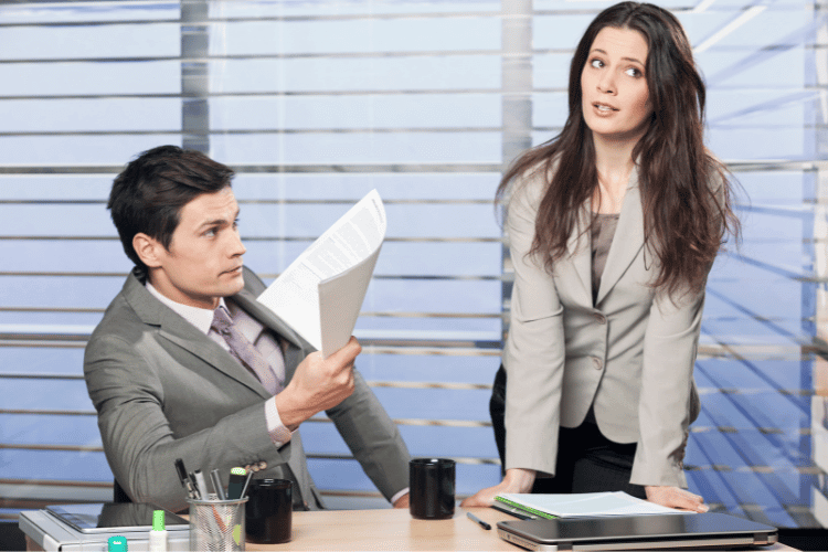 Um homem sentado e uma mulher de pé em um escritório aparentando estarem com um ruído na comunicação.
