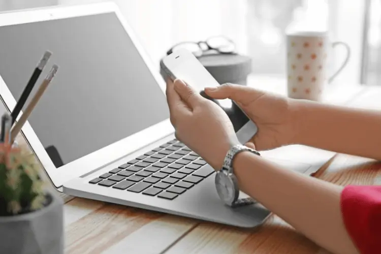 Mãos femininas mexendo em um smartphone e um notebook aberto em sua frente.