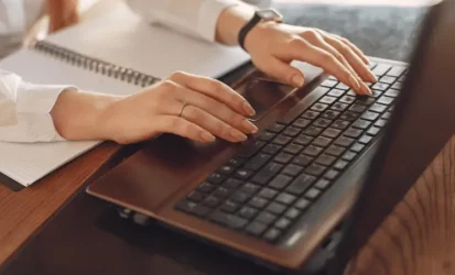 Imagem de mãos femininas digitando em um notebook e um caderno em frente ao computador