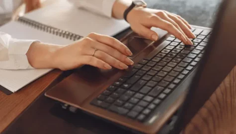 Imagem de mãos femininas digitando em um notebook e um caderno em frente ao computador